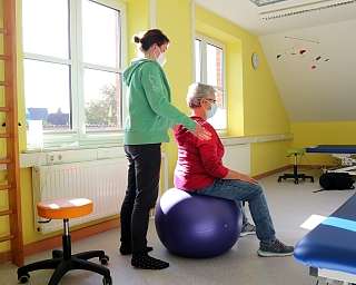 Eine Person sitzt auf einem Gymnastikball, während eine Therapeutin assistiert.